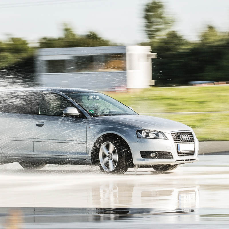 Ein silberner Audi A3 f hrt gischtspritzend mit hoher Geschwindigkeit ber eine nassgespr hte Fahrbahn auf dem Trainingsgel nder das ADAC Fahrsicherheitszentrums. Der Hintergrund ist durch die Geschwindigkeit der mitziehenden Kamera verwischt, man erkenn auf einem grasbewachsenen Wall ein kleines wei es kastenf rmiges Gebaude, dahinter gr ne B ume und Str ucher. Der Himmel ist hellblau, die Szene ist sonnenbeschienen.  Foto: ADAC