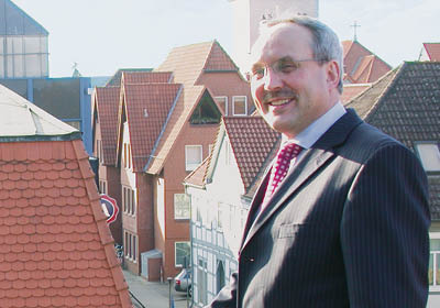 Ein j ngerer J rgen Wache steht auf einem Balkon. Hinter ihm sonnenbeschiene D cher einer Stadt unter blass-blauem Himmel. Foto: Hannoversche Volksbank