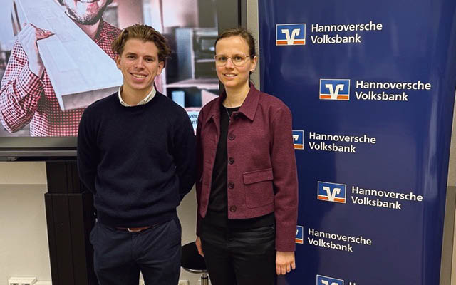 Links neben einem rollup Display stehen links, ein Mann, rechts eine Frau mit Brille in leger–gesch ftlicher Kleidung. Hinter Ihnen ein gro er Monitor mit einem Mann in kariertem Hemd, der eine Holzbowhe auf der Schulter tr gt. Foto: Hannoversche Volksbank
