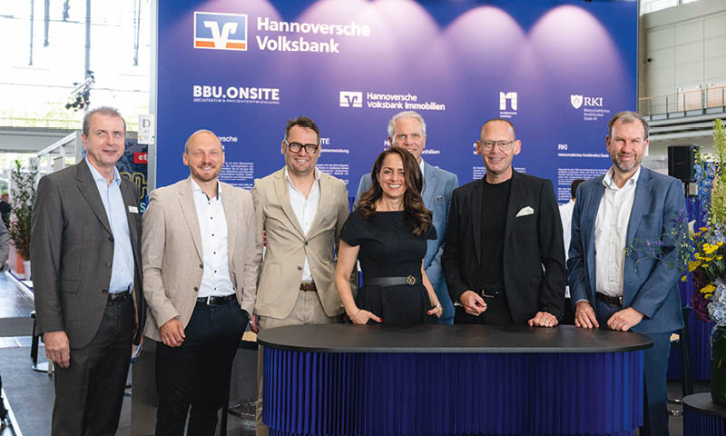 Hinter einem blauen Tresen, vor einem gro en, blauen Leinwand mit dem Logo der Hannoversche Volksbank, oben, links und in kleiner und wei verschiedenen Logos der Volksbank – Unternehmen stehen sechs M nner und eine Frau Business Kleidung.  Foto: Julian Maritz