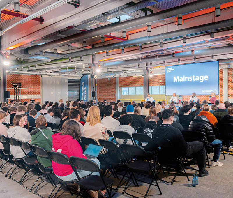 In einer gro en ehemaligen Industriehalle sitzen eine gro e Menge Jugendlicher mit Blick auf eine B hne, auf der vier Referentinnen und Referenten sitzen. Hinter Ihnen eine gro e Leinwand mit der Aufschrift Mainstage. Foto: Alexander D rrer 
