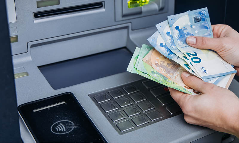 Eine Person vor einem Geldautomaten h lt einige Euro Geldscheine in den H nden. Man sieht nur die H nde mit den Scheinen, und einen Ausschnitt des Geldautomaten im Hintergrund. Im Vordergrund ist ein gro es, leuchtend rotes Warndreieck mit Ausrufezeichen. Foto: wedmoments.stock(shutterstock)