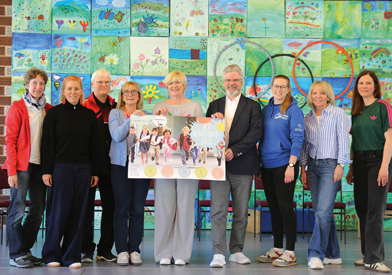 Neun Personen stehen vor einer Wand, die mit vielen bunten Kinderzeichnungen, die kachelartig angeordnet sind, geschm ckt ist. In der Mitte halten drei Personen ein Poster mit dem Bewegungpass-Motiv in die Kamera. Foto: Hannoversche Volksbank