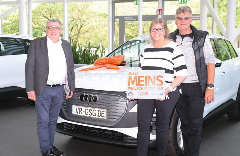 Drei Personen stehen vor einem neuen Audi E-Tron, der eine orangefarbene Schleife auf der vorderen Haube hat. Links Marko Volck, von der Hannoversche Volksbank, rechts die strahlenden Gewinner mit einem Schild in der Hand, auf dem steht: jetzt meins, weil ich mitspiele. Foto: RSB Hannover, Reiner Freese