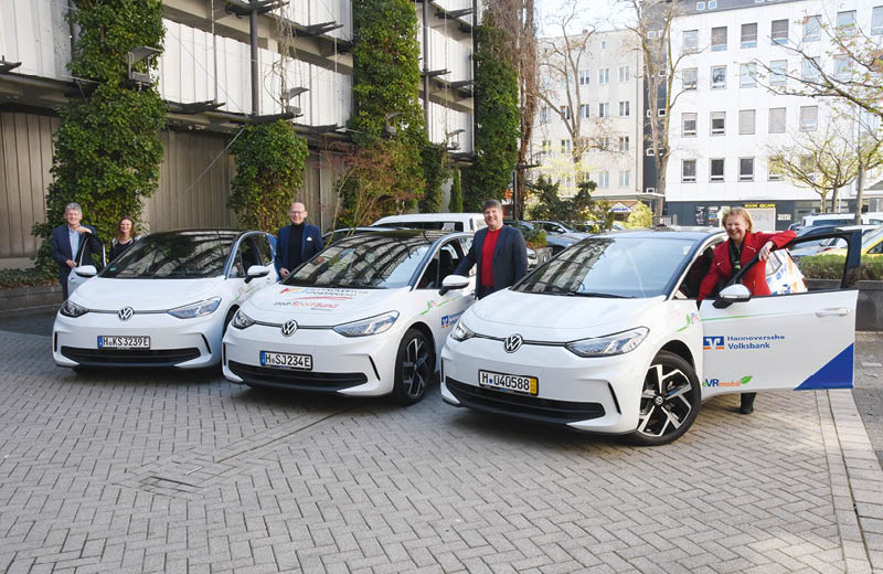 Auf einem Parkplatz stehen drei elektrische VW ID.3, die die Hannoversche Volksbank an drei gemeinn tzige Einrichtungen gespendet hat. Neben dem ge ffneten T ren stehen insgesamt f nf Personen. Im Hintergrund einige B ume, die gerade beginnen zu knospen und mehrst ckige, schmucklose Stadth user. Links die begr nte Betonfassade eines Parkhauses. Foto: Hannoversche Volksbank